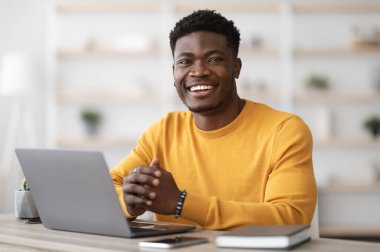 Portrait of positive young black guy in yellow sweater sitting at table, working on laptop, sending emails, office interior. Cheerful african american man freelancer working from home, copy space