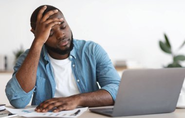 Stresli Siyah Erkek Girişimci 'nin iş yerinde sorunları var. Ofiste dizüstü bilgisayarla üzgün bir şekilde oturan ve umutsuzca kafasını okşayan depresif Afrikalı Amerikalı adam, boş alanla yakın çekim yaptı.