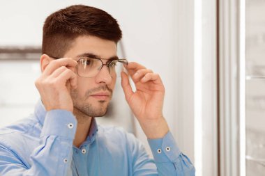 Health Care Concept. Portrait of focused handsome man trying on new glasses in optician store, copy space clipart