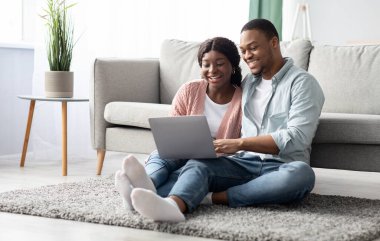 Mutlu siyahi aşıklar halının üzerinde dizüstü bilgisayarlarıyla oturuyor, evde birlikte eğleniyorlar, mekanı kopyalıyorlar. Çevrimiçi mobilya arayan neşeli Afro-Amerikan çift, web sörfü