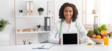 Smiling black lady nutritionist demonstrating digital tablet with empty screen, clinic interior, applications for dieting clipart