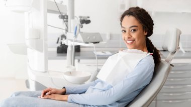 Dişçi koltuğunda oturan ve kameraya gülümseyen genç siyahi bayan.