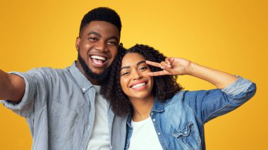 Çift selfie. Neşeli siyahi adam, kız arkadaşıyla fotoğraf çekilirken, kız barış jesti yapıyor, sarı arka planda birlikte poz veriyorlar.
