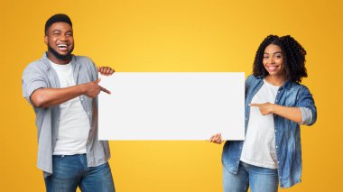 Reklamınız için yer. Neşeli siyah çift ellerinde boş reklam panosuna işaret ediyor, sarı arka planda yan yana duruyorlar, boşluğu kopyalıyorlar.