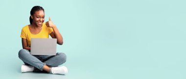 Harika bir site. Siyah kadın dizüstü bilgisayar kullanıyor. Parmak uçlarını yukarı kaldırıyor. Mavi arka planda oturuyor. Stüdyo Çekimi