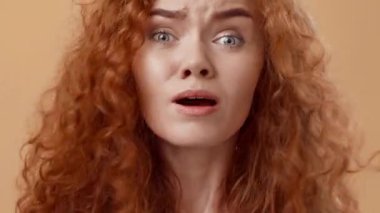 Geniş gözleri ve açık ağzıyla kıvırcık kızıl saçlı genç bir kadın açık bir şaşkınlık ya da şok gösteriyor. Yumuşak bej arka plan onun canlı saç rengini ve duygusal ifadesini arttırır..