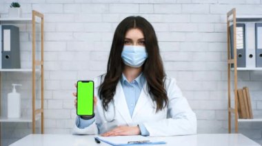 Beyaz önlüklü ve maskeli bir sağlık uzmanı, klinik bir ortamda teknolojiyi ön plana çıkaran boş yeşil ekranlı bir akıllı telefonu gösteriyor..