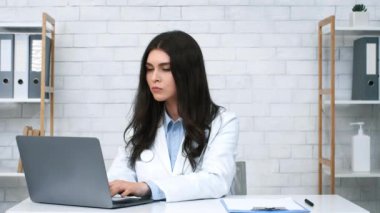 Beyaz önlüklü odaklanmış bir sağlık uzmanı düzenli bir ofis masasında dizüstü bilgisayarda yazıyor. Çevre parlak ve düzenlidir, hasta danışmanlığı ve idari işler için idealdir..