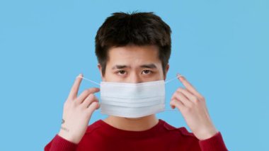 Asyalı genç adam yüzüne tıbbi bir maske takıyor ve takmaya hazırlanıyor. Odaklanmış ve iyi ışıklandırılmış bir alanda gibi görünüyor. Saf mavi bir arkaplanı var. Hareketlerini vurguluyor..