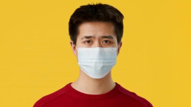 Asyalı genç adam sakince duruyor, açık mavi bir maske takıyor, tarafsız bir ifadeyle. Parlak sarı arka plan kırmızı kazağına zıt düşerek sağlık ve güvenliğe odaklanmasını vurguluyor..