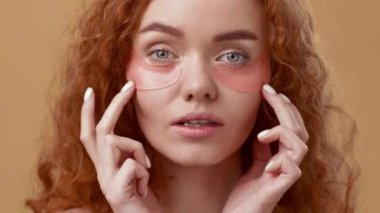 Kıvırcık kızıl saçlı genç bir kadın gözlerinin altına cilt bandı sürerken parlak bir şekilde gülümsüyor. Sıcak arka plan onun neşeli ifadesini ve cilt bakımını arttırıyor..