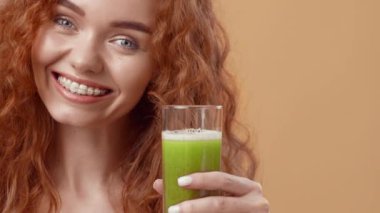 Kıvırcık kızıl saçlı genç bir kadın büyük bir bardak yeşil smoothie tutuyor, sıcak bir şekilde gülümsüyor. Yumuşak, tarafsız arka plan onun neşeli ifadesini arttırıyor, sağlık ve sağlığını vurguluyor..