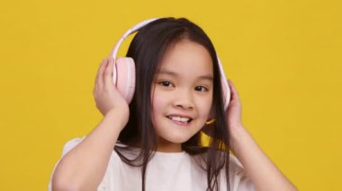 Asyalı kız kulaklıkla müzik dinlemeyi seviyor, canlı sarı bir fon önünde mutlulukla ışıldıyor..