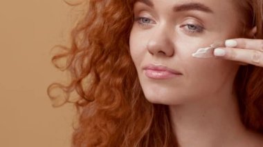 Kıvırcık kızıl saçlı genç bir kadın yüzüne cilt kremi sürerken neşeyle gülümsüyor. Sıcak, nötr arka plan onun ışıl ışıl görünümünü arttırır..