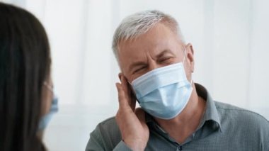 Gri saçlı yaşlı bir adam, sağlık ofisinde maske takarken bir kadınla sağlık sorunlarını tartışıyor. Atmosfer profesyonel ve hasta bakımına odaklı..