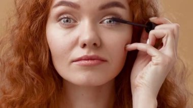 Kıvırcık saçlı genç bir kadın yavaşça aydınlatılmış bir odada otururken dikkatle dikkatle odaklanan kirpiklerine rimel sürer. Sıcak tonlar güzelliğini ve özelliklerini arttırıyor..