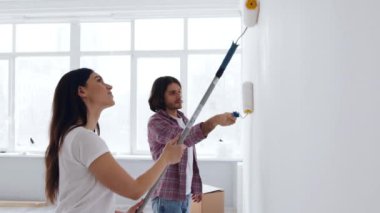 Bir çift oturma odasının duvarını boyayarak DIY projesinin tadını çıkarıyor. Odayı dolduran doğal ışıkla çevrili boya ruloları ve kahkahalar kullanıyorlar..