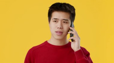 Kırmızı kazak giyen Asyalı genç adam telefonunda odaklanmış bir ifadeyle konuşuyor. Canlı sarı arkaplan o ana enerji katıyor. Bu da gündelik bir konuşma olduğunu gösteriyor..