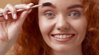 Kıvırcık saçlı neşeli genç bir kadın kaşlarını evde şekillendirmek için fırça kullanıyor. Güneş ışığı yüzünü aydınlatıyor, parlak gülüşünü ve doğal güzelliğini vurguluyor..
