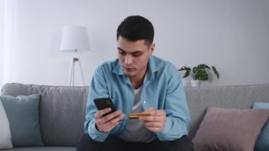Genç bir adam kanepeye oturmuş elinde kredi kartıyla akıllı telefonuna odaklanmış. Oturma odasında bir lamba ve saksı bitkisi var, rahat bir atmosfer yaratıyor..