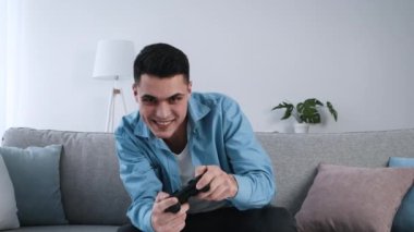 Genç bir adam kanepede rahatça oturur ve elinde bir kumandayla oyun oynamaya odaklanır. Modern oturma odasında hafif ışıklandırma ve ev bitkileri var. Rahatlatıcı bir atmosfer yaratıyorlar..