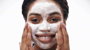 Afro-Amerikalı bir kadın gülümserken yüzüne kremsi bir maske takar ve kendine bakmanın tadını çıkarır. Parlak arka plan onun parıldayan cildini ve rahatlamış davranışlarını vurguluyor..