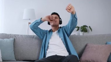 Genç bir adam gri bir kanepeye oturur, kulaklık takar ve müzik dinlerken neşeyle dans eder. Rahat oturma odasında hafif ışıklandırma ve iç mekan bitkileri var..