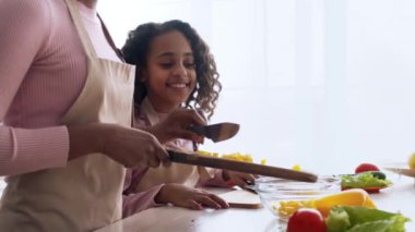 Siyah anne ve kızı birlikte yemek pişiriyorlar. Annesi lezzetli bir salata için ustalıkla sebze doğrarken kız gülümsüyor. Neşeli anlar parlak mutfakta yakalanır..