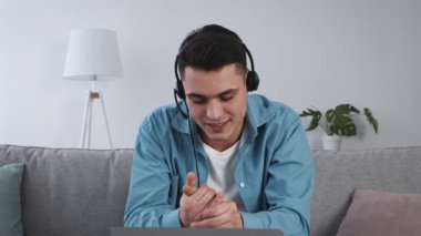 Genç bir adam sanal bir toplantıya odaklanmış, kulaklık ve günlük kıyafetler giyiyor. Parlak bir oturma odasındaki kanepeye rahatça oturur ve tartışmaya aktif olarak katılır..