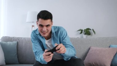 Genç bir adam rahat bir kanepede video oyunları oynamaktan zevk alır. Oyun kontrolörüne odaklanırken gülümser. Parlak oturma odasında yumuşak mobilyalar ve yakınlarda bir bitki var..