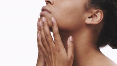 Afro-Amerikalı kadın cilt bakımı yaparken yüzüne hafifçe dokunuyor, parlak, minimalist bir ortamda sakin bir ifade sergiliyor. Yumuşak ışık, onun doğal güzelliğini ve huzurunu vurgular..