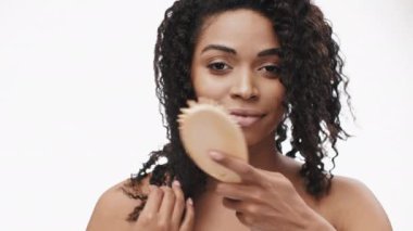 Afro-Amerikalı neşeli genç kadın doğal güzelliğini ve özgüvenini sergileyerek kıvırcık saçlarını tarıyor. Aydınlatıcı ve davetkar bir ortam. Mutluluğunu vurguluyor..