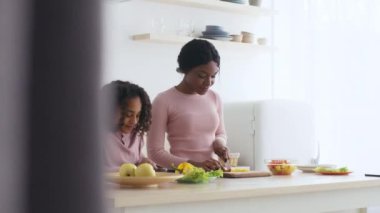 Kızı yanında otururken Afrikalı Amerikalı bir kadın mutfak tezgahında meyve kesiyor. Modern beyaz bir mutfaktalar..