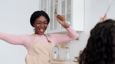 Pembe bluzlu ve önlüklü mutlu bir siyah kadın, parlak bir mutfakta kıvırcık saçlarını şekillendirirken dans ediyor. Atmosfer neşeli ve canlı, yaratıcılık ve neşe dolu..