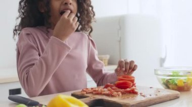 Genç siyah bir kız, ahşap bir kesim tahtasına sebze yerleştirirken bir domates parçasını ısırıyor. Parlak mutfağında sağlıklı bir yemek hazırlamanın hazzına dalmış..