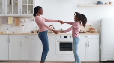 Neşeli siyah bir anne ve kız, samimi mutfaklarında el ele dans ediyor, birlikte güzel vakit geçiriyorlar. Odada beyaz dolaplar ve sıcak ahşap aksanlar var. Ev atmosferi yaratıyor..