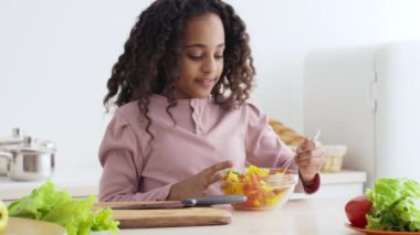 Bir kız, etrafını marul ve mutfak malzemeleriyle çevirmiş, gülümserken bir kasede taze meyve karıştırıyor. Doğal ışık, neşeli ve davetkar mutfak alanını aydınlatır..