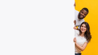 Beyaz reklam panosunun arkasından bakan mutlu ırklar arası çift. Mesajınız için boş yer var, sarı stüdyo arka planı.