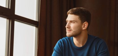 Millennial Guy Portresi. Kendine güvenen genç adam, modern çatı katında, pencerenin yanında oturuyor. Boşluğu Kopyala