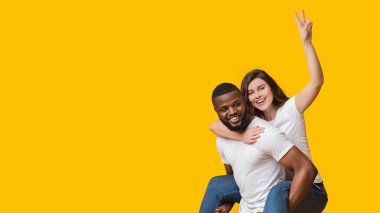 Afro erkek arkadaşlarına binen neşeli bir kadın barış jesti ya da V işareti göstererek sarı arka planda birlikte eğleniyor.