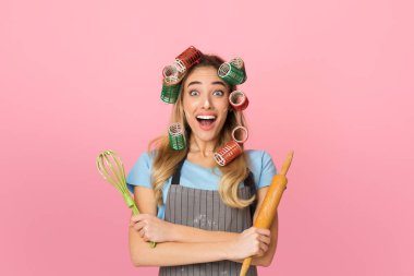 Surprised housewife with curlers opens mouth wide with shock, holding whisk and wooden rolling pin, isolated over pink background