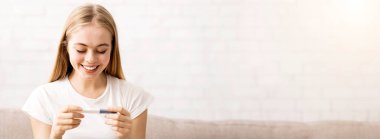 Happy young blonde woman looking at pregnancy test in her hands, home interior, panorama