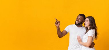 Şuraya bak. Sarı arkaplan ve panorama üzerindeki fotokopi alanına sarılıp işaret eden neşeli çok ırklı çift.