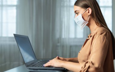 Sosyal Mesafe. Yüz maskesi takan, dizüstü bilgisayar kullanan, tahta masada oturan bir kızın yan görüntüsü.