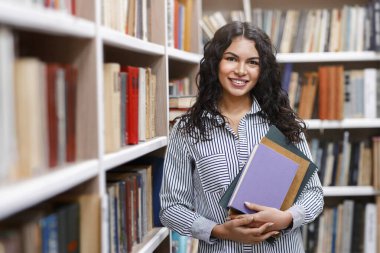 Akademik kavram. Mutlu gülümseyen Latin kadın elinde kitaplarla kampüs kütüphanesinde kitap raflarına yaslanıyor.