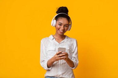 Müzik Uygulaması. Akıllı telefonlu ve kablosuz kulaklıklı mutlu afro kız en sevdiği şarkıları dinliyor ve kamera, sarı arka plan