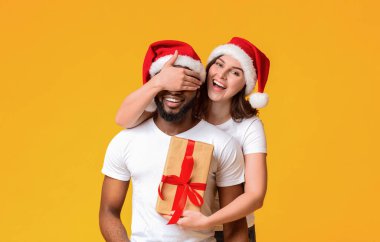 Noel Baba şapkalı neşeli genç bir kadın, siyah erkek arkadaşına Noel hediyesi ile sürpriz yapıyor, sarı stüdyo arka planında birlikte poz veriyor.