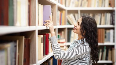 Genç Kütüphaneci. Gülümseyen Meksikalı kız kütüphane kitaplığından kitap alıyor, el kitabını arıyor ve seçiyor.
