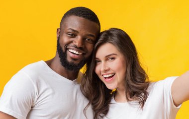 Mutlu ırklar arası çift selfie çekiyor, içtenlikle gülümsüyor ve sarı arka planda birlikte poz veriyorlar.
