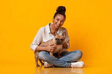 Beautiful Afro Woman Posing With Her Cute French Bulldog Puppy, Sitting With Crossed Legs On Floor Over Yellow Background In Studio, Copy Space